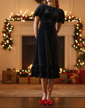 Woman in a black dress with bows standing in a festive room with Christmas decorations.
