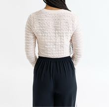 Person wearing a textured beige long-sleeve top and dark pants on a white background